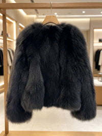 A dark gray to black faux fur jacket hanging on a wooden hanger on a clothing rack inside a boutique.