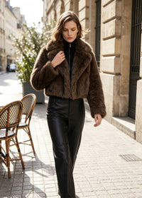 A woman walking on a sunlit city sidewalk wearing a cropped brown faux-fur jacket over a black top and black leather pants.
