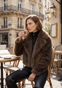 Lifestyle street style shot of a woman drinking espresso at an outdoor café wearing a cozy cropped espresso brown faux fur jacket.