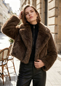 A woman wearing a brown faux fur cropped jacket over a black top and black leather pants, standing on a city street.
