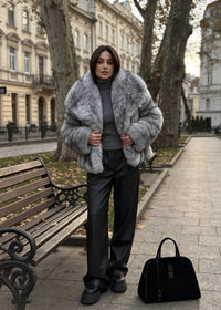 Woman standing in an autumn park modeling an open front gray faux fur coat styled with black pants and a matching black tote bag