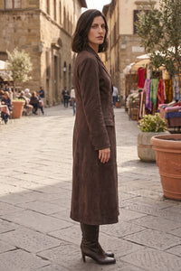A woman stands in profile on a cobblestone street wearing a long brown suede-like coat and dark heeled boots, with market stalls and buildings in the background.