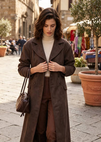 A woman stands on a cobblestone street wearing a long brown double-breasted coat over a cream turtleneck, paired with brown trousers and carrying a brown shoulder bag.
