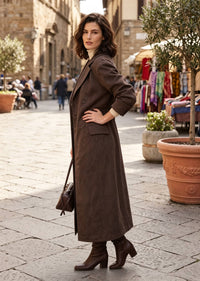 A woman stands on a cobblestone street wearing a long brown coat over a cream turtleneck, paired with brown ankle boots and carrying a small brown handbag. The background shows a blurred outdoor market scene with stone buildings and potted plants.