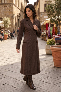 A woman wearing a long brown double-breasted coat and matching brown boots, walking down a cobblestone street in an outdoor town setting with market stalls and buildings in the background.