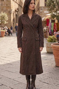 A woman stands in a stone street wearing a long brown suede-like double-breasted coat with wide lapels and front flap pockets, paired with dark brown knee-high boots.