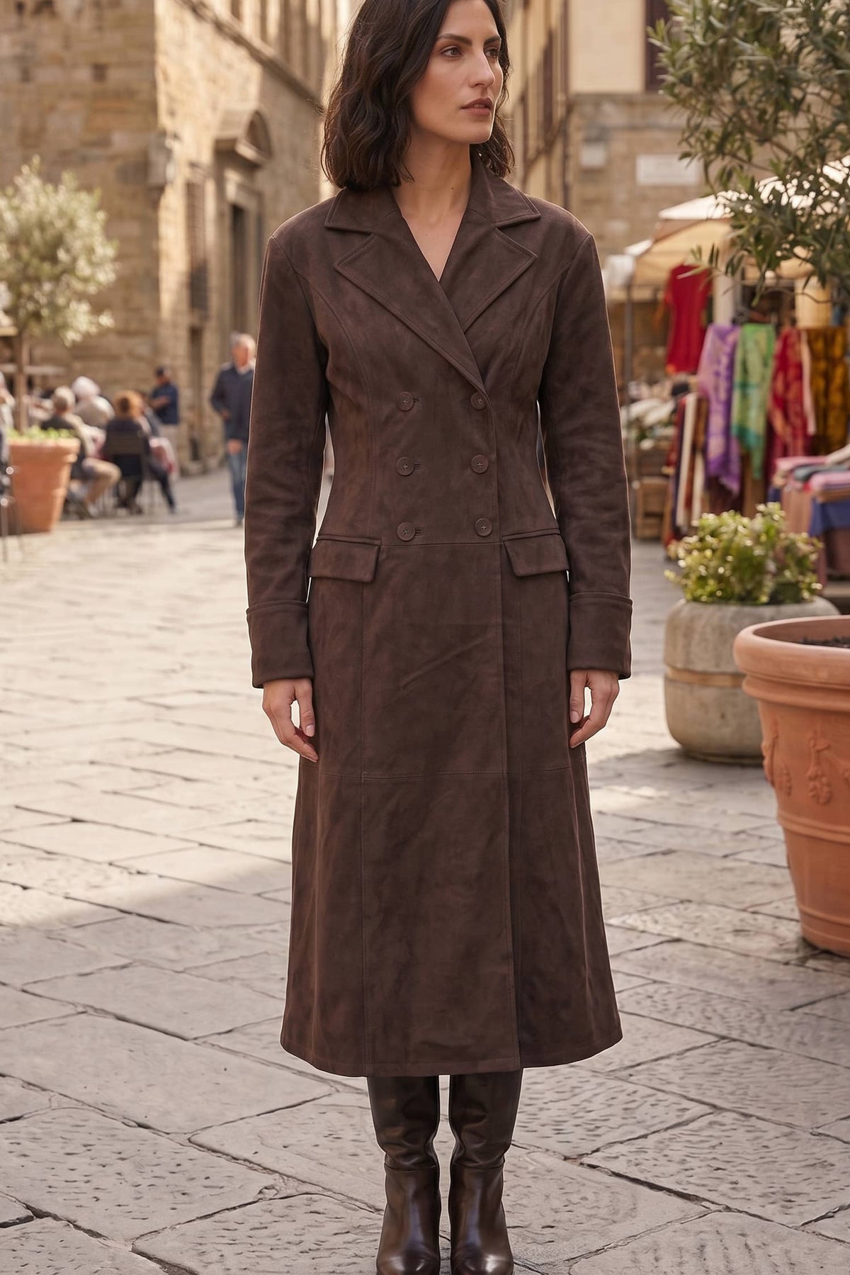 A woman stands in a stone street wearing a long brown suede-like double-breasted coat with wide lapels and front flap pockets, paired with dark brown knee-high boots.