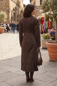 A woman viewed from behind wearing a long brown suede-like coat and carrying a matching brown handbag, standing on a stone-paved street in an outdoor market setting.
