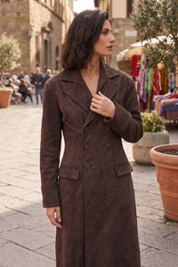 A woman wearing a long, double-breasted brown suede-like coat stands in an outdoor street setting with stone buildings and potted plants in the background.