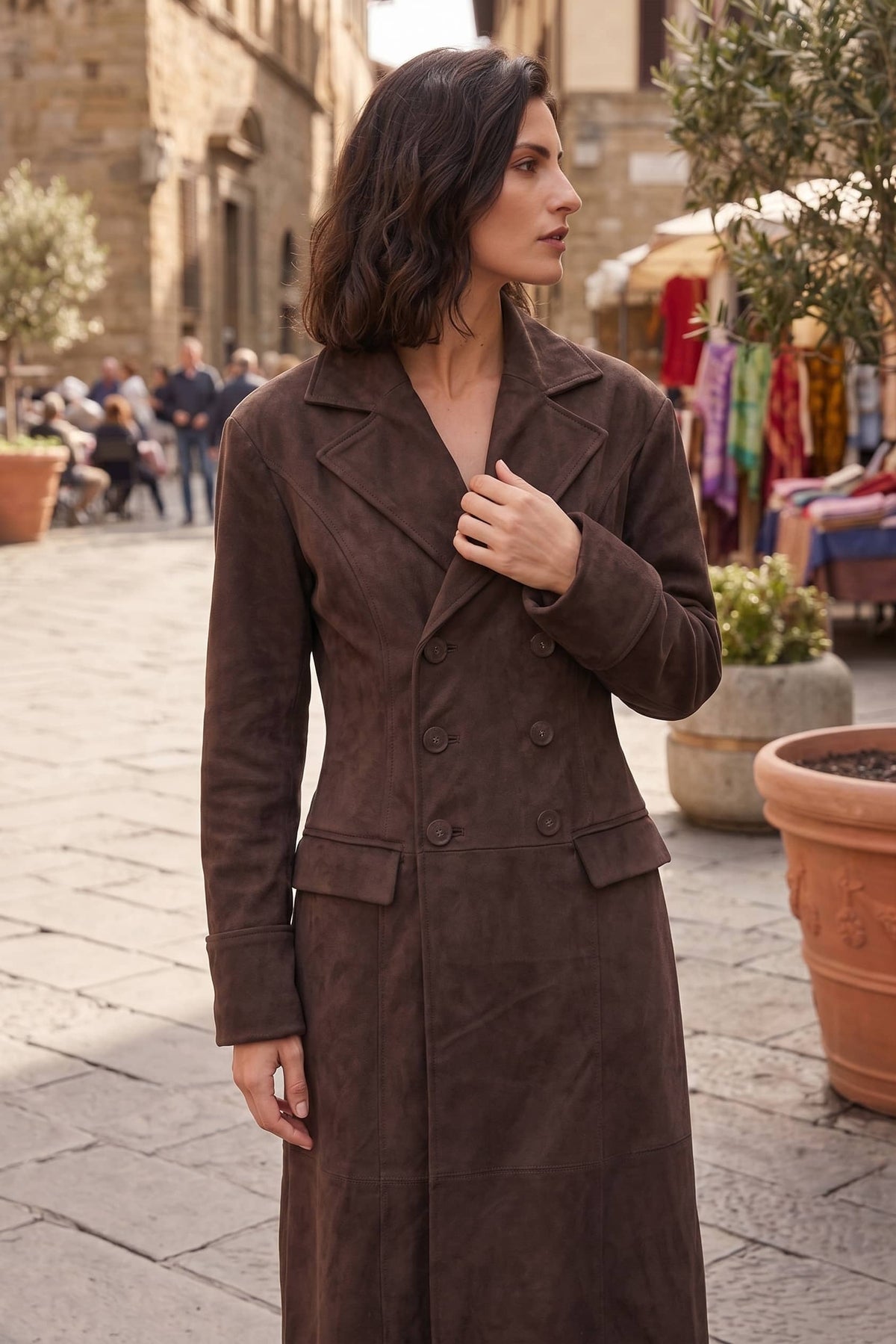 A woman wearing a long, double-breasted brown suede-like coat stands in an outdoor street setting with stone buildings and potted plants in the background.