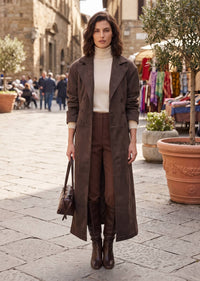 A woman stands on a cobblestone street wearing a long brown suede-like trench coat over a cream turtleneck, brown trousers, and knee-high brown boots, while carrying a brown handbag.