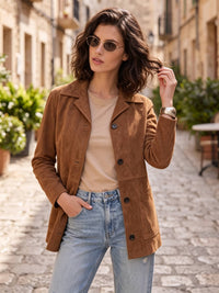 A woman wearing a brown suede jacket over a beige top and light blue jeans, standing on a cobblestone street in a European-style town.