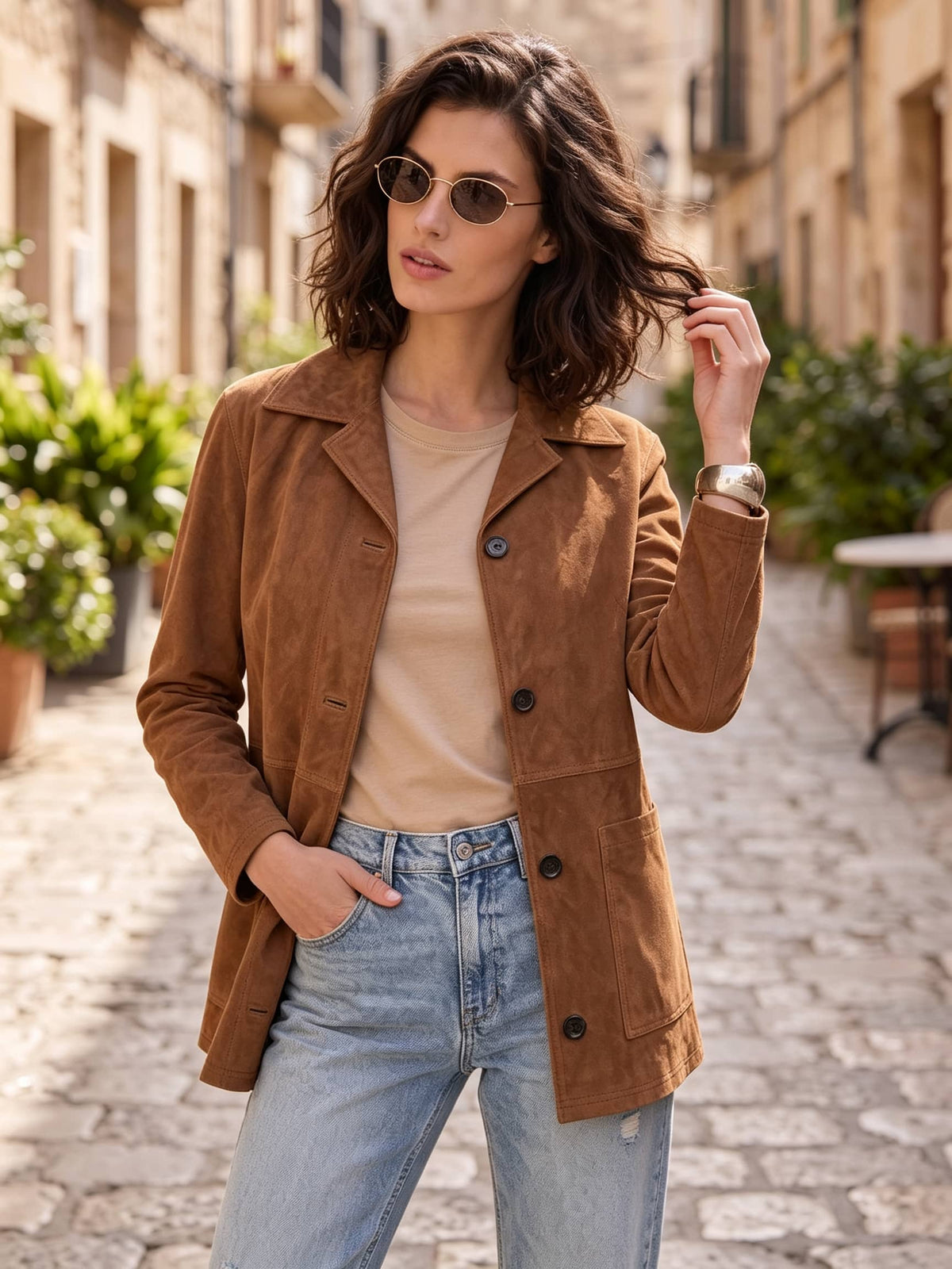 A woman wearing a brown suede jacket over a beige top and light blue jeans, standing on a cobblestone street in a European-style town.