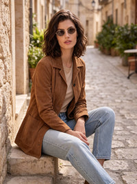 A woman wearing a brown suede jacket, beige t-shirt, and light blue jeans sits on stone steps in a sunny, narrow European street.