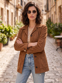 A woman wearing a brown suede jacket, light blue jeans, and sunglasses stands with arms crossed on a cobblestone street in a European-style town.