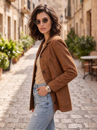 A woman wearing a brown suede blazer, beige top, and light blue jeans stands on a cobblestone street in a European-style town, wearing round sunglasses.