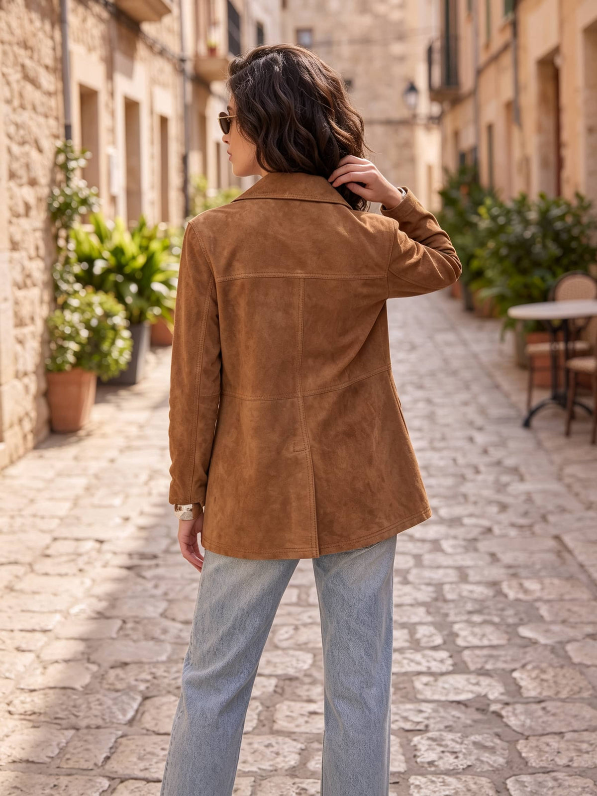 A woman wearing a brown suede blazer and light blue jeans, photographed from behind in a sunlit cobblestone street lined with stone buildings and potted plants.