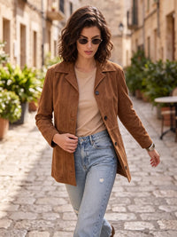 A woman walking down a cobblestone street wearing a brown suede jacket over a beige top and light blue jeans.