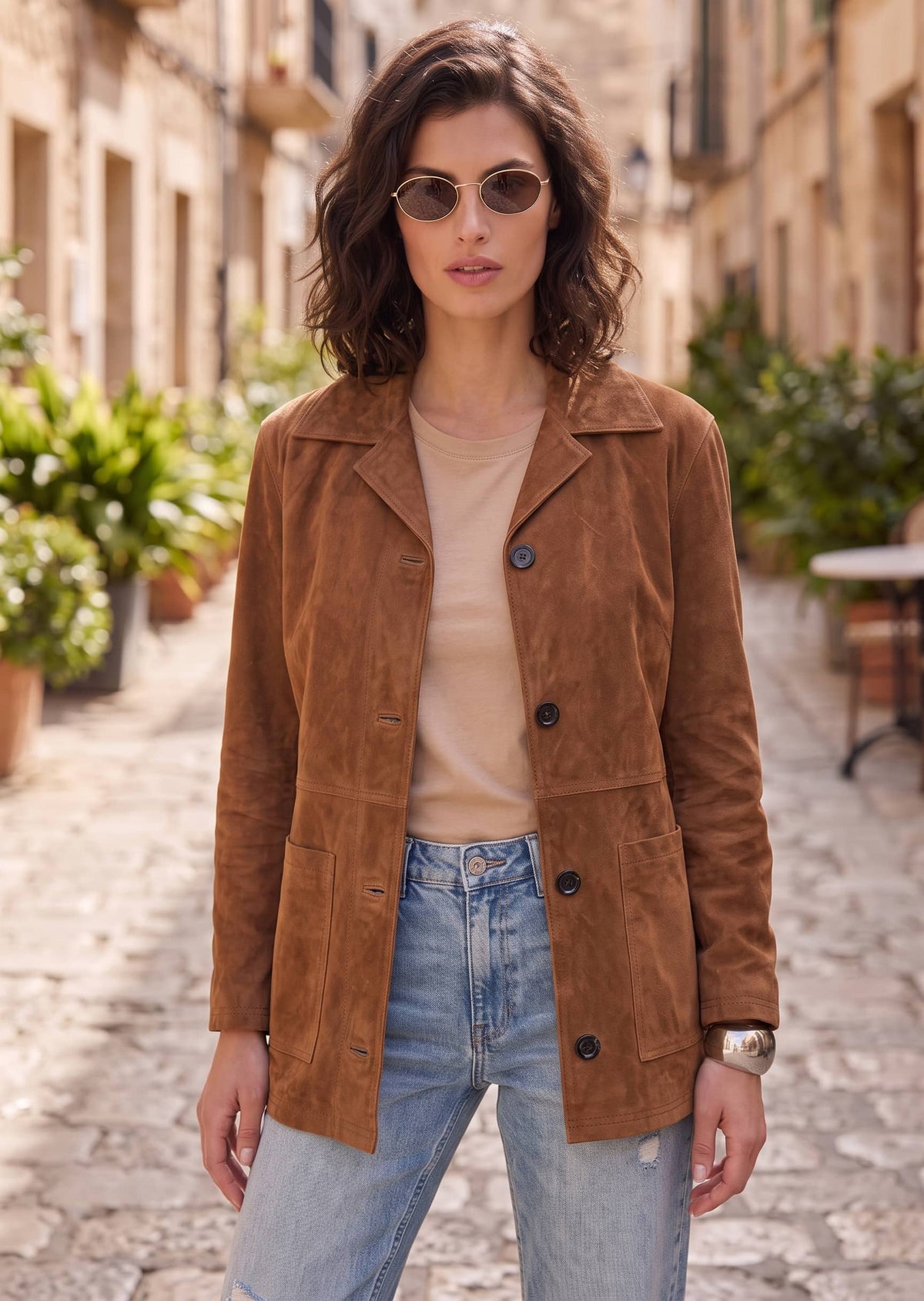 A woman wearing a brown suede jacket over a beige top and light blue jeans, standing on a cobblestone street in a European-style town.