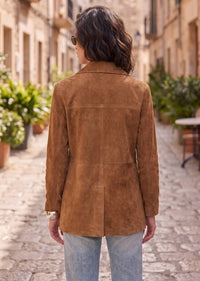 A woman viewed from behind wearing a tan suede jacket and light blue jeans, standing on a cobblestone street in a European-style town with buildings and potted plants in the background.