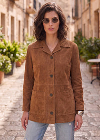 A woman wearing a brown suede jacket and light blue jeans stands on a cobblestone street in a European-style town, wearing round sunglasses.
