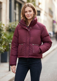 A woman wearing a short, quilted puffer jacket in a deep burgundy or maroon color, paired with dark blue jeans, standing on a blurred city street.