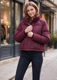 A woman wearing a cropped, quilted puffer jacket in a deep burgundy/maroon color, paired with dark blue jeans, standing on a city street.
