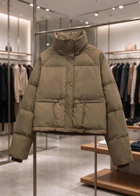A light brown puffer jacket with a high stand-up collar and zippered front, hanging on a display stand inside a modern clothing store.