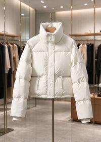 A white puffer jacket with a high stand-up collar and front snap closure, displayed on a hanger on a dress form stand inside a modern clothing boutique.