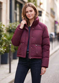 A woman wearing a cropped, quilted puffer jacket in a deep burgundy/maroon color, standing on a blurred city street.