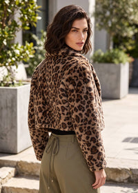 A woman wearing a cropped faux-fur jacket with a brown and black leopard print, paired with olive green drawstring pants, photographed outdoors in a sunny urban setting.