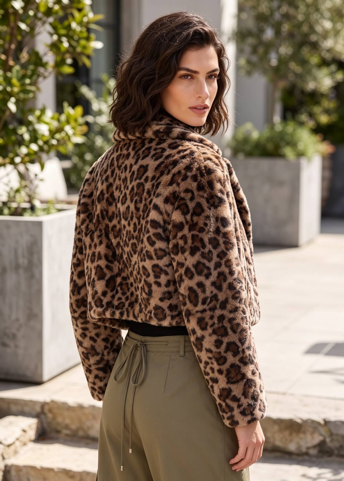A woman wearing a cropped faux-fur jacket with a brown and black leopard print, paired with olive green drawstring pants, photographed outdoors in a sunny urban setting.