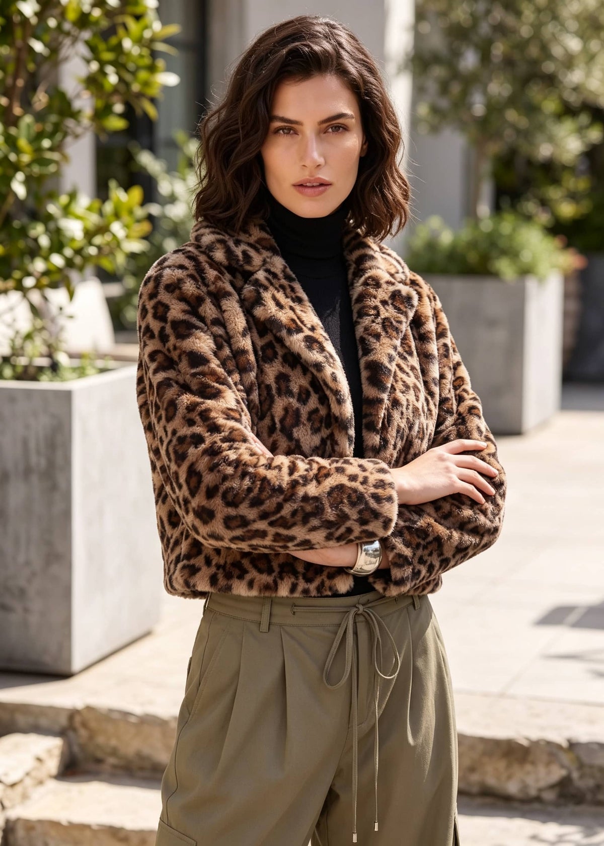 A woman wearing a brown and black leopard print faux fur jacket over a black turtleneck and olive green drawstring pants, standing outdoors in front of a modern building with stone steps and planters.