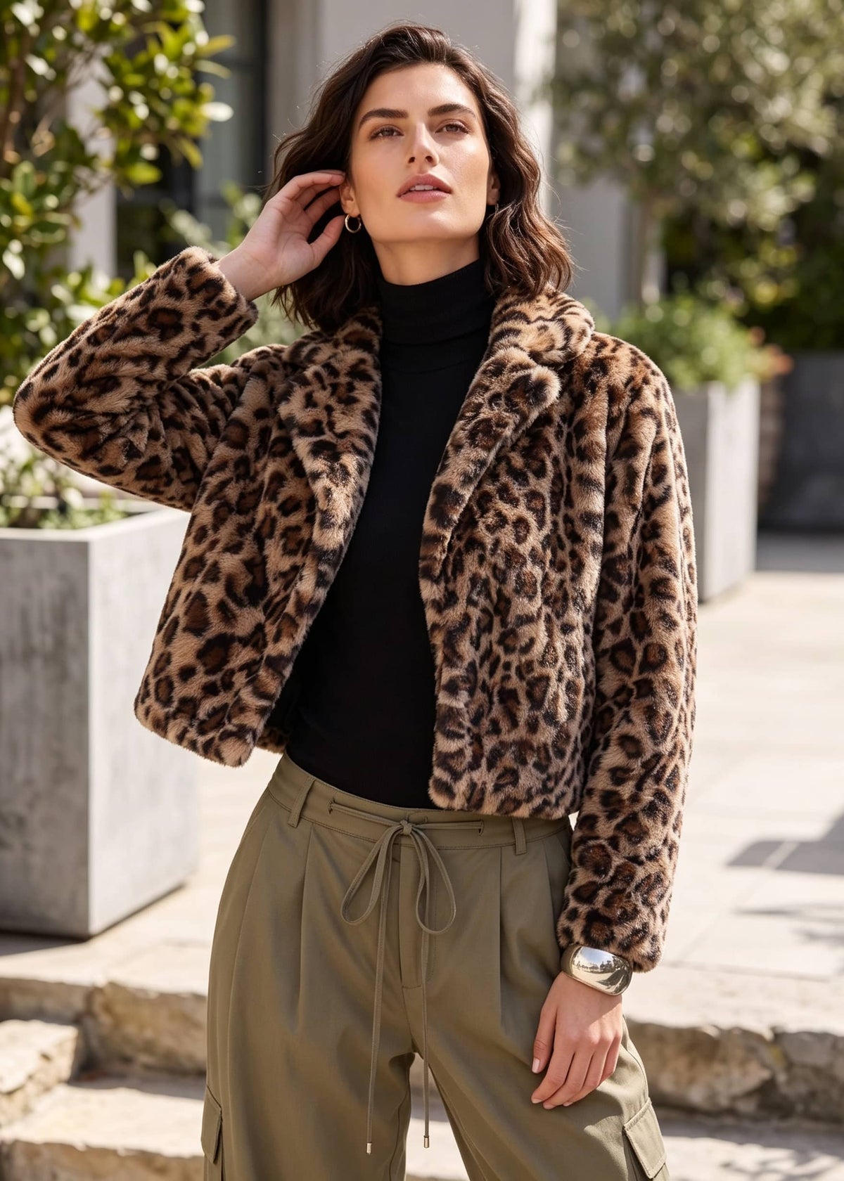 A woman wearing a plush leopard-print faux fur jacket over a black turtleneck and olive green drawstring pants, standing outdoors on stone steps.