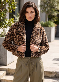 A woman wearing a cropped leopard-print faux fur jacket over a black turtleneck and olive green drawstring pants, standing outdoors in front of a modern building.