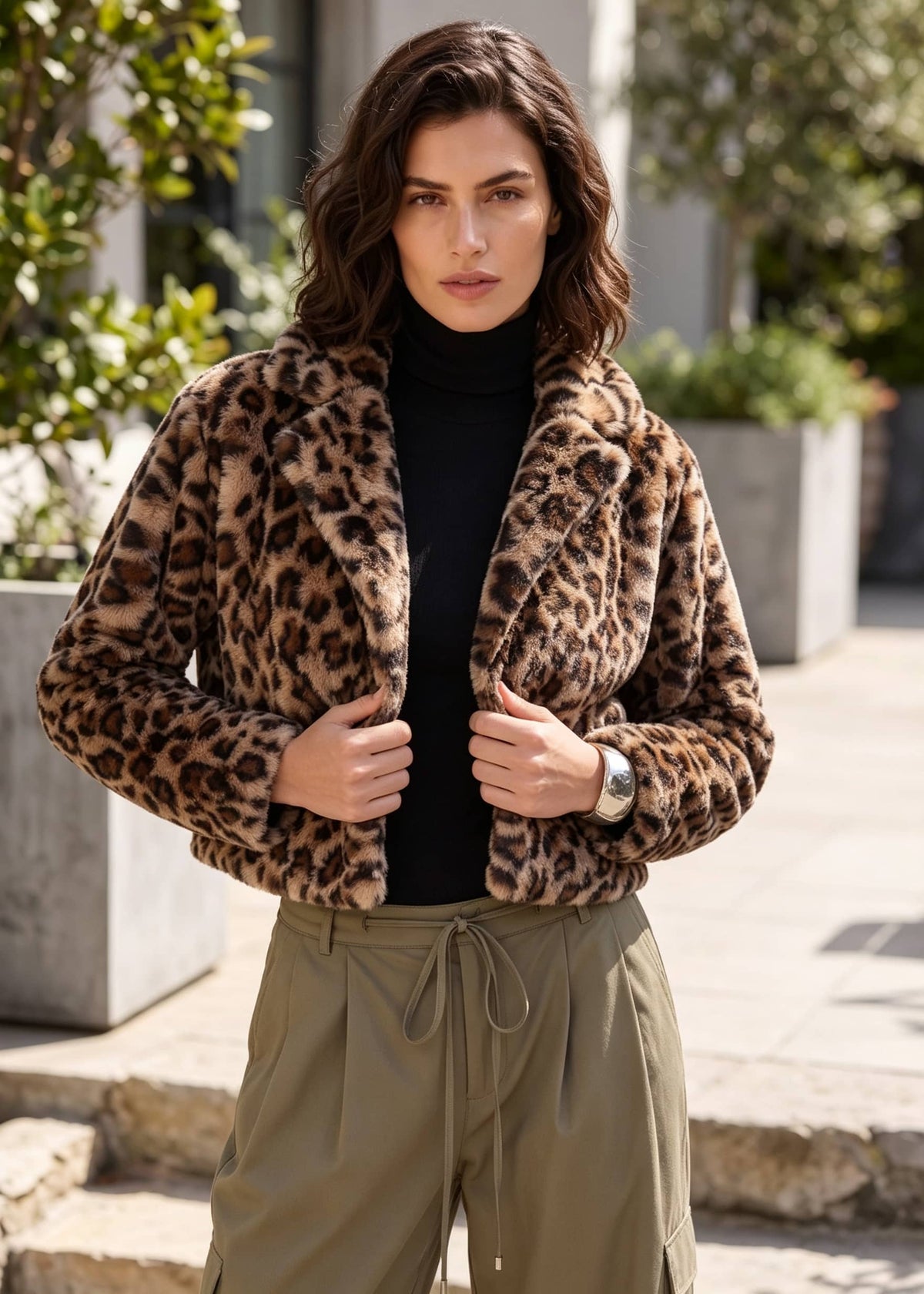 A woman wearing a cropped leopard-print faux fur jacket over a black turtleneck and olive green drawstring pants, standing outdoors in front of a modern building.