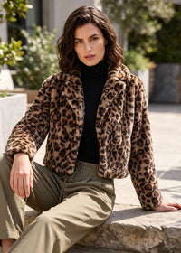 A woman wearing a cropped leopard-print faux fur jacket over a black turtleneck and olive green pants, sitting on a stone ledge in an outdoor patio setting.