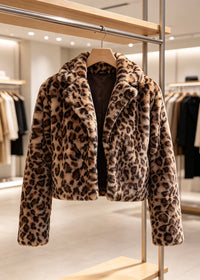 A cropped faux fur jacket with a brown and beige leopard print pattern, hanging on a wooden hanger on a clothing display stand inside a boutique.