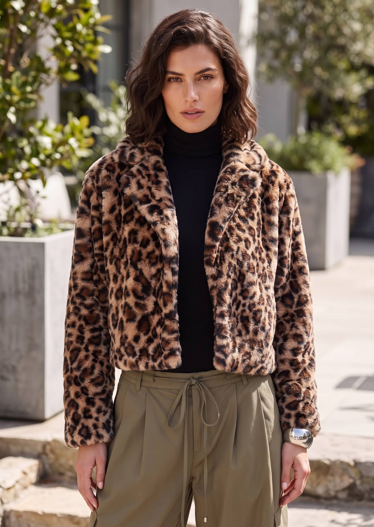 A woman wearing a cropped leopard-print faux fur jacket over a black turtleneck and olive green drawstring pants, standing outdoors in front of a modern building with greenery.