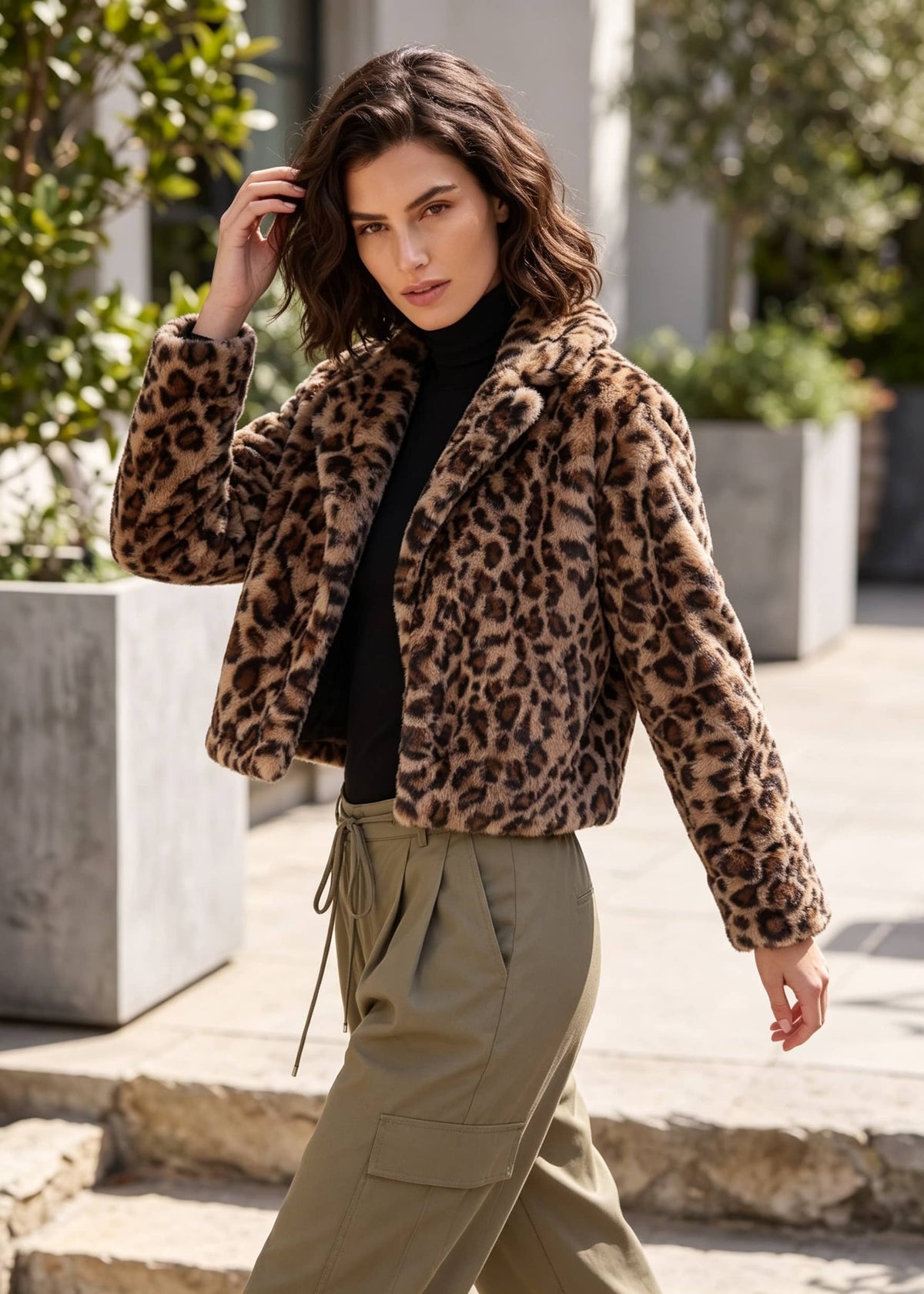 A woman wearing a cropped leopard-print faux fur jacket over a black turtleneck and olive green cargo pants, walking outdoors on stone steps.