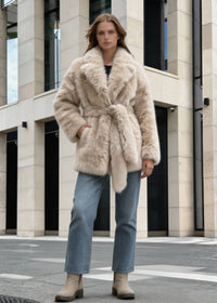 Full-length front view of a woman wearing a cream belted faux fox fur wrap coat, blue jeans, and beige boots in front of a modern building