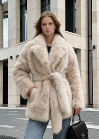 Close-up of a model posing in a plush cream belted faux fur jacket holding a black quilted handbag.