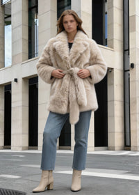 Full-body shot of a woman adjusting the waist belt of a beige faux fur wrap coat with wide lapels.