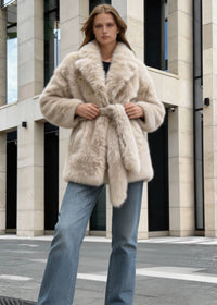  Woman tying the self-tie belt on a luxury beige faux fur coat in an urban street setting.