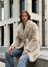 Seated view of a woman wearing a cozy plush beige faux fox fur coat outdoors.