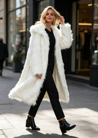 Blonde woman walking past city shops wearing a long white faux fur coat with oversized notch lapels, black trousers, and studded ankle boots.