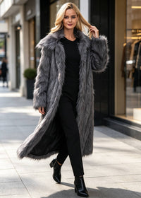 Dynamic walking view of a smiling blonde woman wearing an open charcoal grey longline faux fur winter coat on an urban sidewalk.