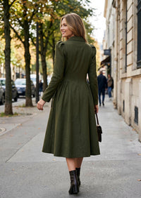 A woman wearing an olive green long-sleeve midi dress with structured shoulders and a flared skirt, walking on a city sidewalk. She is carrying a small dark handbag and wearing dark brown ankle boots.