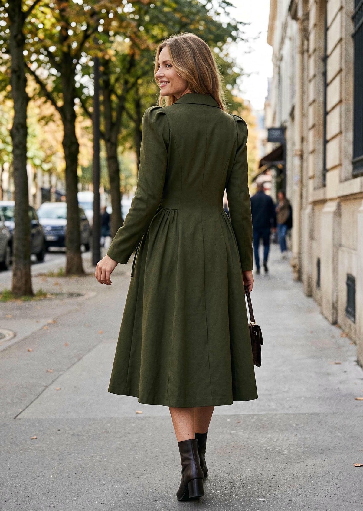 A woman wearing an olive green long-sleeve midi dress with structured shoulders and a flared skirt, walking on a city sidewalk. She is carrying a small dark handbag and wearing dark brown ankle boots.