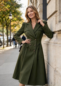 A woman wearing an olive green double-breasted trench coat leans against a stone building on a city sidewalk.
